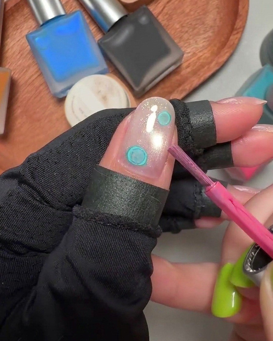 Ein Pinsel fügt rosa Aquarell-Tintenpunkte neben bestehenden blauen Punkten auf einem gelbeschichteten Fingernagel hinzu.