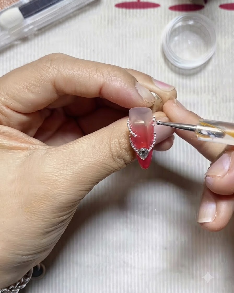 Ein Dotting-Tool platziert einzelne silberne Stahlperlen entlang einer geschwungenen Klebelinie auf einem verzierten Fingernagel.